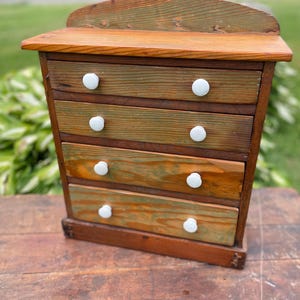 May include: A small wooden chest of drawers featuring four horizontal drawers with visible wood grain and green-toned staining. Each drawer is equipped with two small, round, white knobs. The unit has a flat top shelf and a curved backboard.