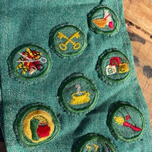 Op de afbeelding: Close-up van een groene denim stof versierd met diverse geborduurde patches. De patches tonen kleurrijke ontwerpen, waaronder sleutels, een koffiekopje en andere gedetailleerde illustraties. De patches zijn cirkelvormig met groene randen.