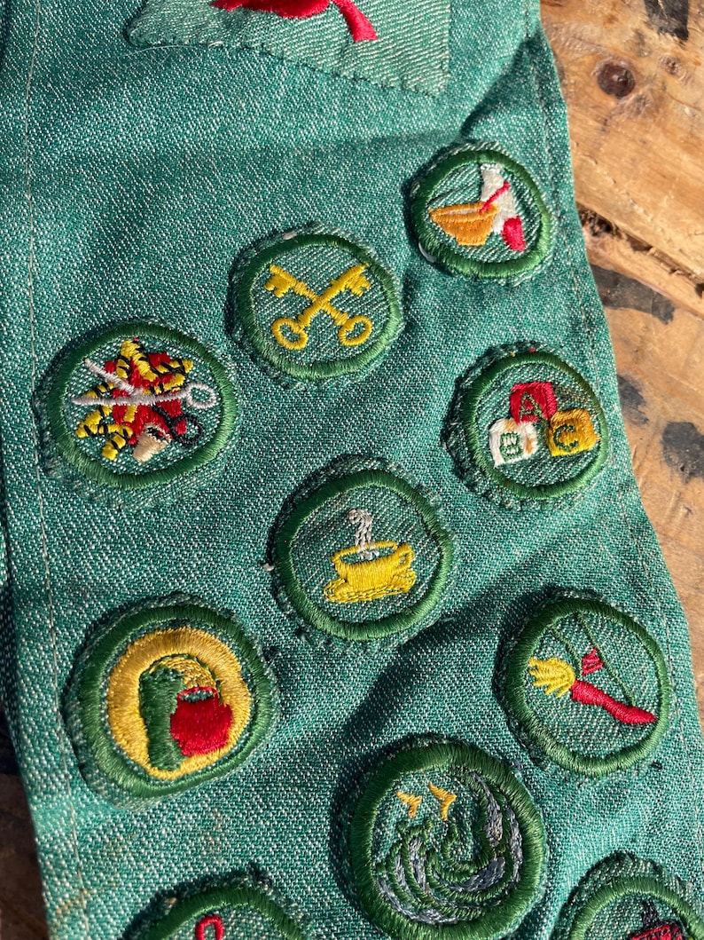 Op de afbeelding: Close-up van een groene denim stof versierd met diverse geborduurde patches. De patches tonen kleurrijke ontwerpen, waaronder sleutels, een koffiekopje en andere gedetailleerde illustraties. De patches zijn cirkelvormig met groene randen.