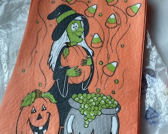 May include: A rectangular orange paper napkin featuring a cartoon illustration of a green-skinned witch holding a pumpkin. A jack-o'-lantern sits beside a black cauldron filled with green bubbles, while candy corn shapes float in the background.