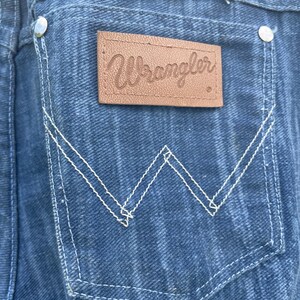 Pu&ograve; includere: Primo piano di una tasca di un capo in denim con cuciture bianche e una toppa in pelle marrone. La toppa presenta il nome del marchio "Wrangler" in corsivo. Il denim &egrave; di colore blu medio.