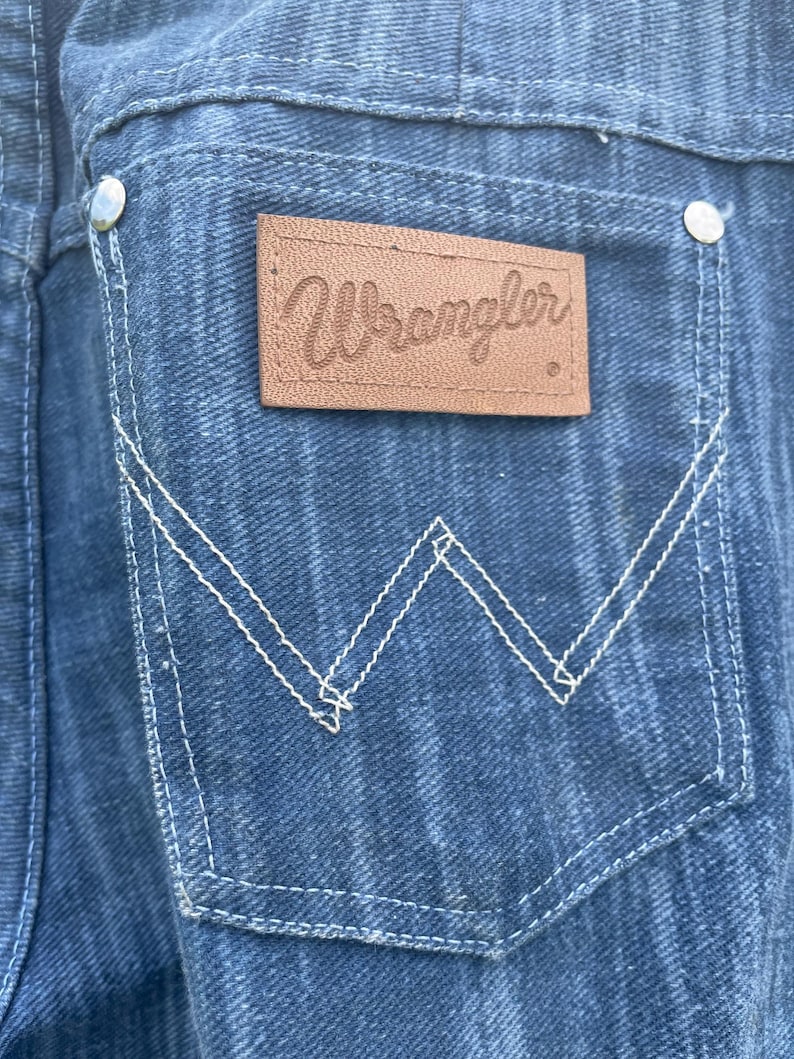 Pu&ograve; includere: Primo piano di una tasca di un capo in denim con cuciture bianche e una toppa in pelle marrone. La toppa presenta il nome del marchio "Wrangler" in corsivo. Il denim &egrave; di colore blu medio.