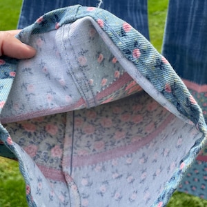 Pu&ograve; includere: Primo piano dell'orlo di un paio di jeans blu. La fodera interna presenta un motivo floreale con rose rosa su sfondo azzurro. Una striscia rosa borda il tessuto floreale.