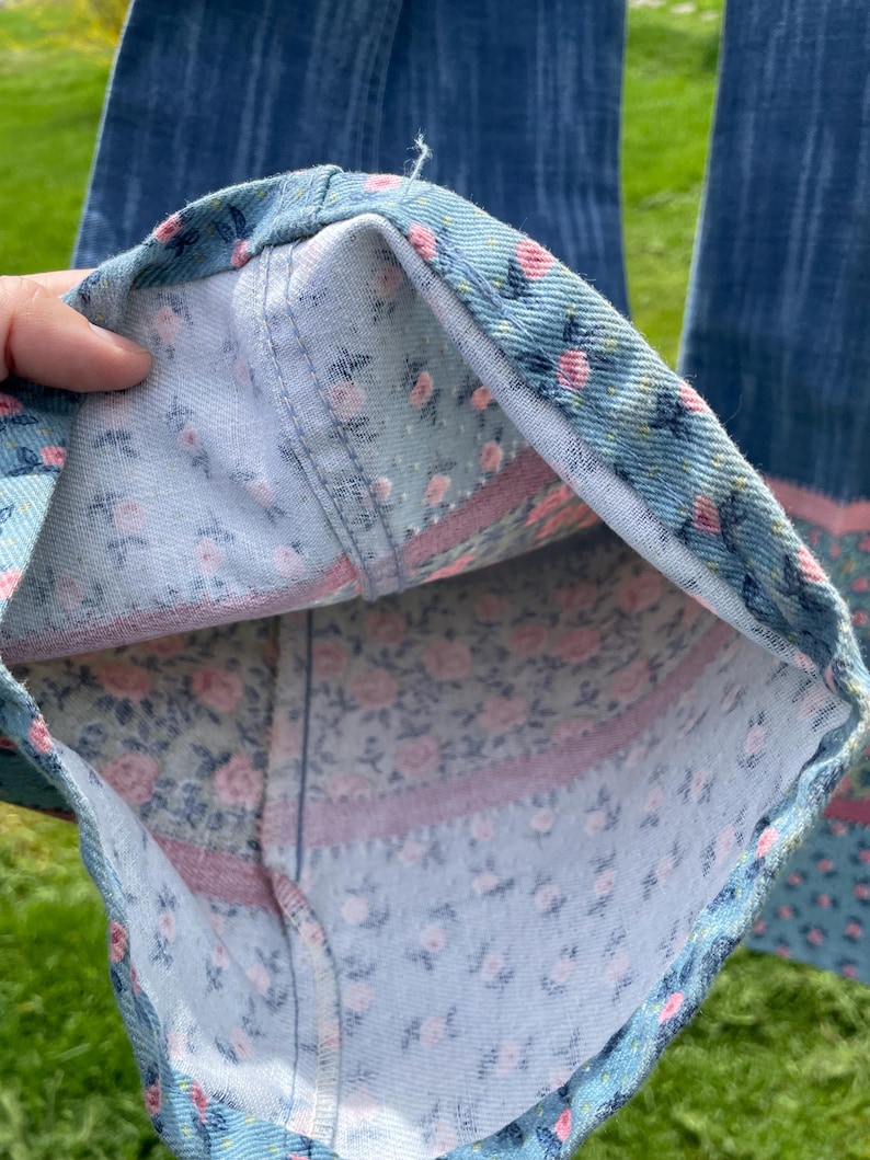Pu&ograve; includere: Primo piano dell'orlo di un paio di jeans blu. La fodera interna presenta un motivo floreale con rose rosa su sfondo azzurro. Una striscia rosa borda il tessuto floreale.