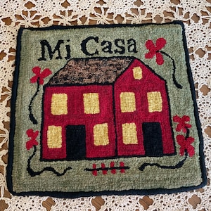 May include: A square, hand-stitched rug with a red house design and the words "Mi Casa" in black. The house has yellow windows and a brown roof, set against a green background with red flower accents. The rug is bordered in black.