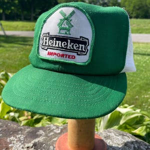 May include: A green and white trucker hat featuring a white patch on the front with a green windmill graphic, the black text Heineken, and the red text IMPORTED. The cap has a solid green front panel, a green brim, and a white mesh back panel.