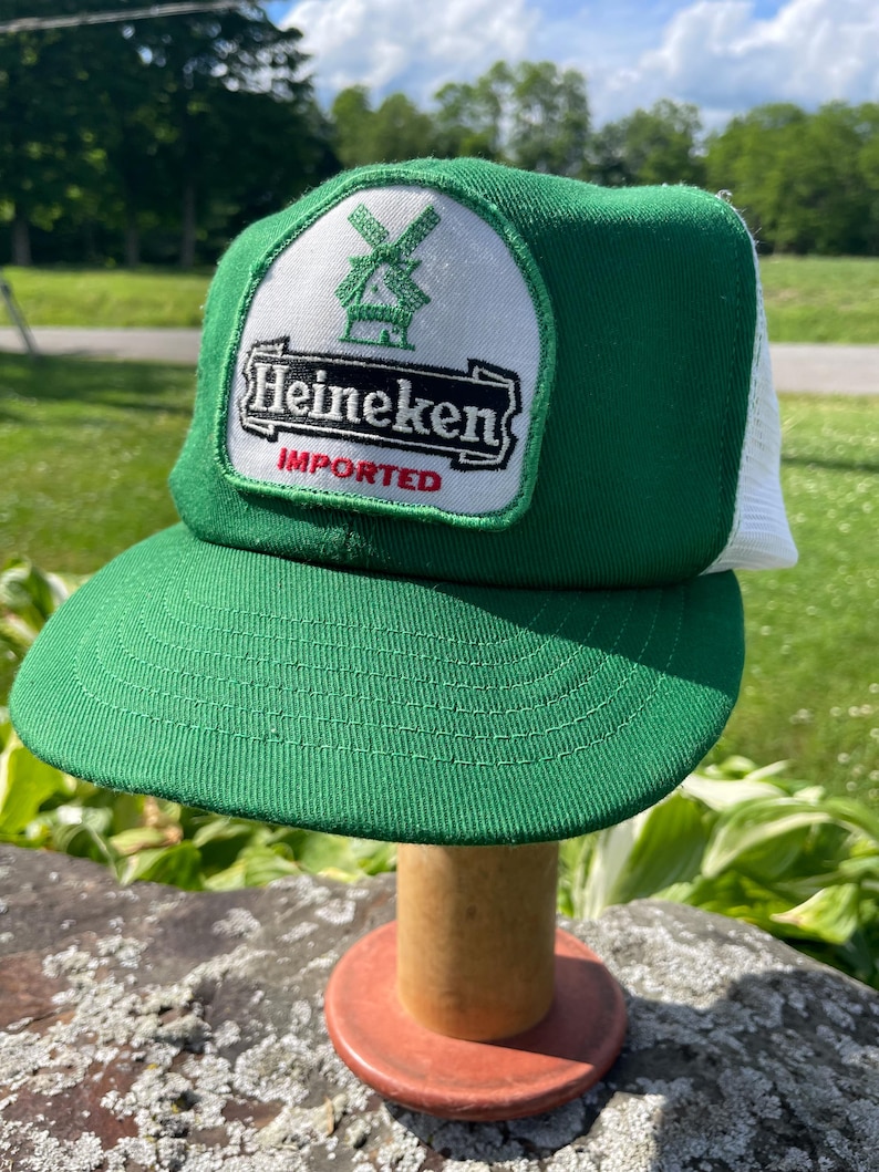 May include: A green and white trucker hat featuring a white patch on the front with a green windmill graphic, the black text Heineken, and the red text IMPORTED. The cap has a solid green front panel, a green brim, and a white mesh back panel.