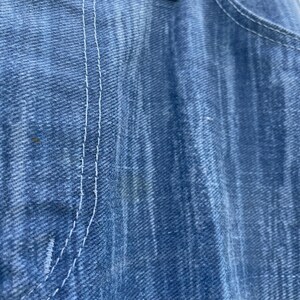 Pu&ograve; includere: Primo piano di jeans di denim blu, che mostra la trama del tessuto e i dettagli delle cuciture bianche. L'immagine evidenzia la tasca e la chiusura con bottoni dei jeans. I jeans sembrano usurati.