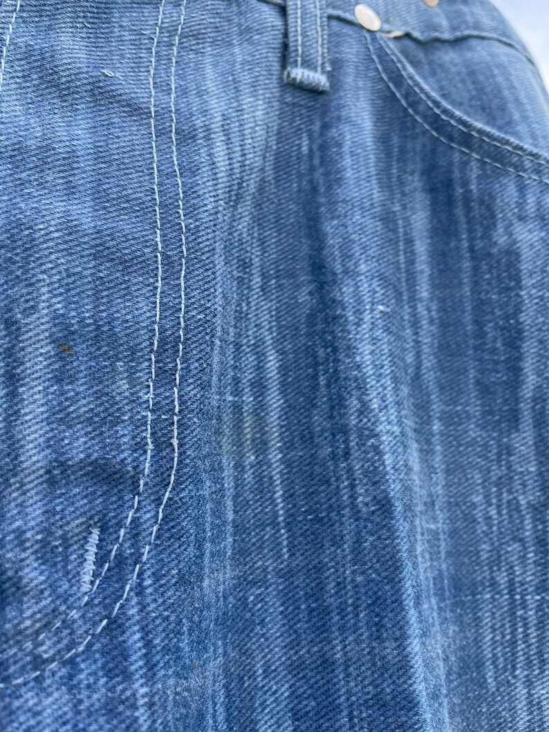 Pu&ograve; includere: Primo piano di jeans di denim blu, che mostra la trama del tessuto e i dettagli delle cuciture bianche. L'immagine evidenzia la tasca e la chiusura con bottoni dei jeans. I jeans sembrano usurati.
