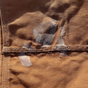 May include: Close-up of a section of brown work pants, showing a pocket and stitching details. Patches of dried, light-colored material are visible on the fabric, suggesting wear and use. The texture of the heavy-duty fabric is clearly visible.
