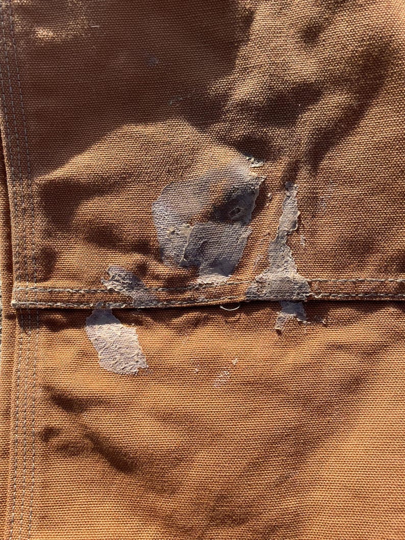 May include: Close-up of a section of brown work pants, showing a pocket and stitching details. Patches of dried, light-colored material are visible on the fabric, suggesting wear and use. The texture of the heavy-duty fabric is clearly visible.