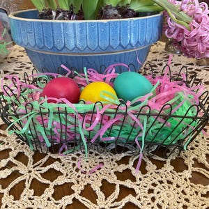 May include: A decorative metal basket filled with colorful Easter eggs and shredded paper. The basket contains a red, yellow, and teal egg, nestled in green and pink paper. A blue ceramic pot with a plant and pink flowers are in the background.