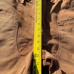 May include: Close-up of brown work pants with a yellow measuring tape. The tape reads up to 16 inches. The pants have pockets and metal rivets. The text on the tape reads "Ellis Bros. Fine Home Furnishings" and "159-161 WASHINGTON S".