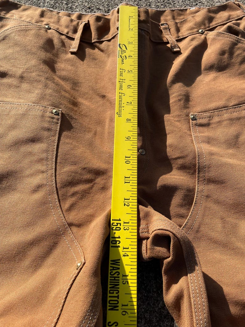 May include: Close-up of brown work pants with a yellow measuring tape. The tape reads up to 16 inches. The pants have pockets and metal rivets. The text on the tape reads "Ellis Bros. Fine Home Furnishings" and "159-161 WASHINGTON S".