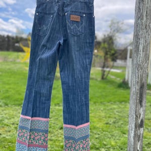 Pu&ograve; includere: Jeans Wrangler blu con orlo floreale. I jeans sono appesi a un filo per stendere i panni. Il motivo floreale presenta fiori rosa e verdi su sfondo blu. Il logo Wrangler &egrave; visibile sulla tasca posteriore.