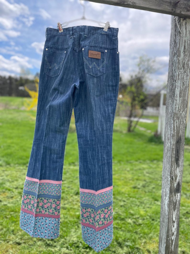 Pu&ograve; includere: Jeans Wrangler blu con orlo floreale. I jeans sono appesi a un filo per stendere i panni. Il motivo floreale presenta fiori rosa e verdi su sfondo blu. Il logo Wrangler &egrave; visibile sulla tasca posteriore.