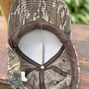 Op de afbeelding: Een baseballpet met een camouflagepatroon in bruin-, groen- en zwarttinten. De binnenkant toont een wit voorpaneel, bruine stoffen voering, een metalen verstelgesp met de opdruk Made in USA en een Mossy Oak-logo op de klep.