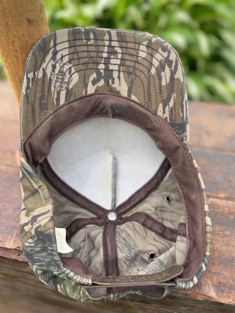Op de afbeelding: Een baseballpet met een camouflagepatroon in bruin-, groen- en zwarttinten. De binnenkant toont een wit voorpaneel, bruine stoffen voering, een metalen verstelgesp met de opdruk Made in USA en een Mossy Oak-logo op de klep.