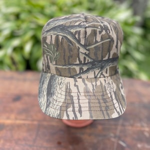 Op de afbeelding: Een baseballpet met een bruin, beige en zwart camouflagepatroon met een takontwerp. De pet heeft een gestructureerde kroon en een gebogen klep met zichtbare stiksels. Een klein rond logo is op de rechterkant van de klep gedrukt.