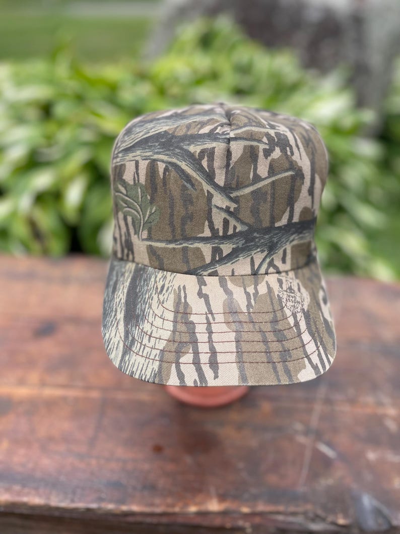 Op de afbeelding: Een baseballpet met een bruin, beige en zwart camouflagepatroon met een takontwerp. De pet heeft een gestructureerde kroon en een gebogen klep met zichtbare stiksels. Een klein rond logo is op de rechterkant van de klep gedrukt.