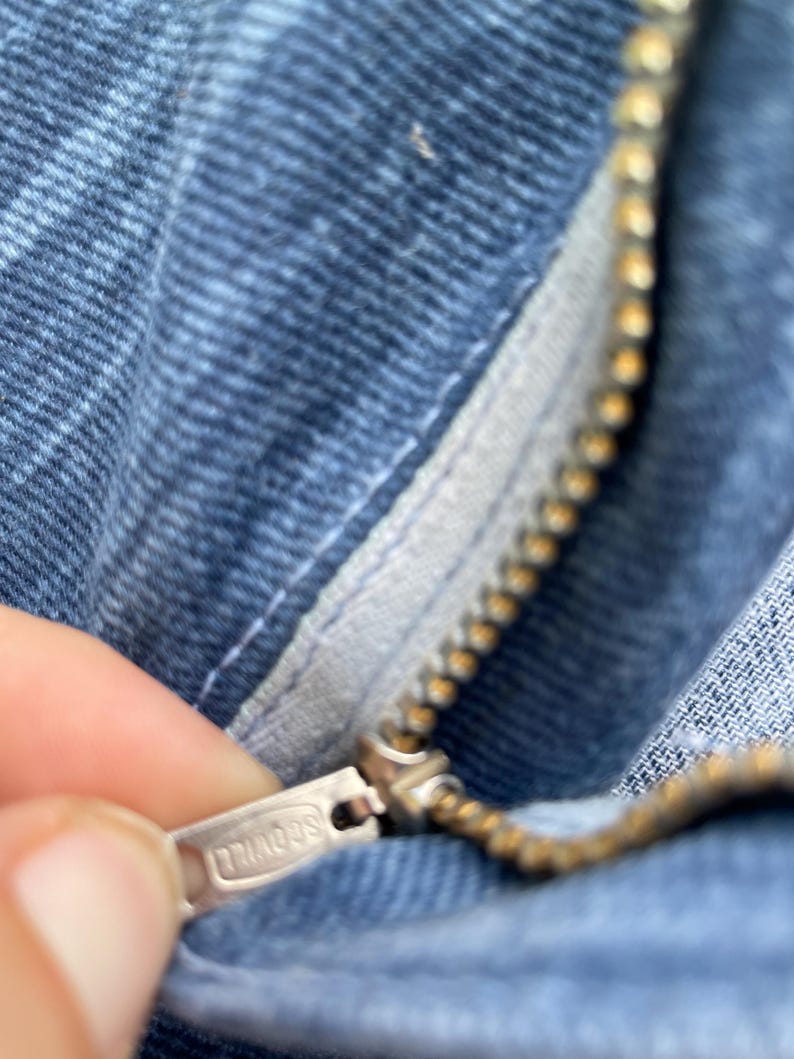 Pu&ograve; includere: Primo piano della cerniera di jeans blu. La cerniera &egrave; color oro e il tirante &egrave; argentato con la scritta "TRIADOS" incisa. Il tessuto denim &egrave; un lavaggio blu medio.