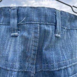 Pu&ograve; includere: Primo piano di jeans di denim blu, che mostrano la cintura, i passanti e i dettagli delle cuciture. I jeans sono appesi a una gruccia di metallo su uno sfondo di cielo nuvoloso. Le cuciture sono bianche.