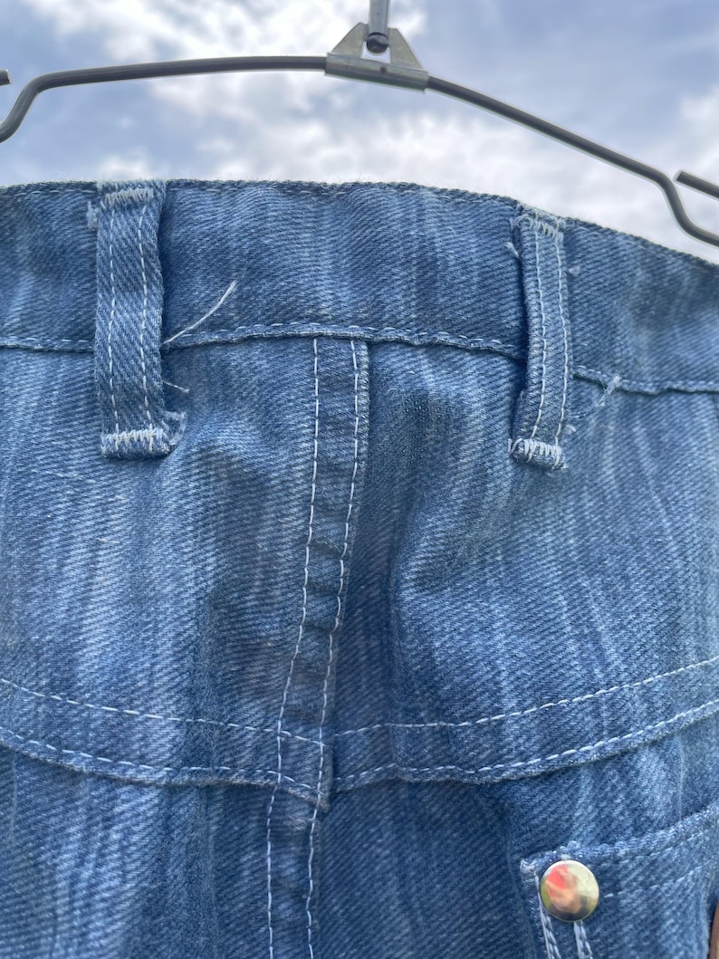 Pu&ograve; includere: Primo piano di jeans di denim blu, che mostrano la cintura, i passanti e i dettagli delle cuciture. I jeans sono appesi a una gruccia di metallo su uno sfondo di cielo nuvoloso. Le cuciture sono bianche.