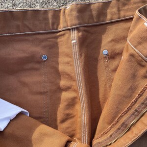 May include: Close-up of brown work pants with white stitching and metal rivets. The pants have a zipper closure and a white fabric peeking out from the inside. The fabric appears to be a durable, heavy-duty material.