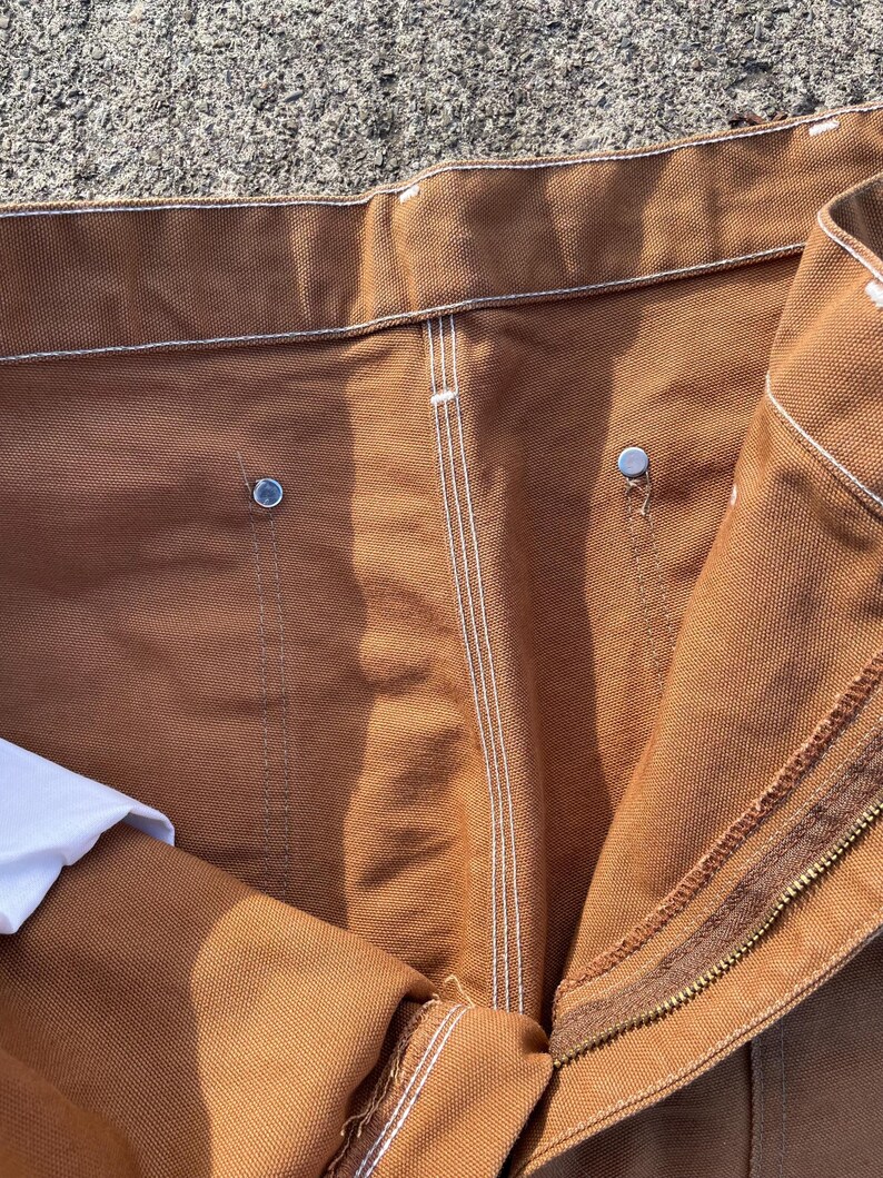 May include: Close-up of brown work pants with white stitching and metal rivets. The pants have a zipper closure and a white fabric peeking out from the inside. The fabric appears to be a durable, heavy-duty material.