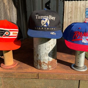 May include: Three baseball caps on display stands. The cap on the left is red with black and white stripes and a Flyers logo. The middle cap is black with a blue brim and a Tampa Bay Lightning logo. The cap on the right is blue with a red brim and a New York Rangers logo.