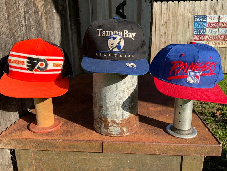 May include: Three baseball caps on display stands. The cap on the left is red with black and white stripes and a Flyers logo. The middle cap is black with a blue brim and a Tampa Bay Lightning logo. The cap on the right is blue with a red brim and a New York Rangers logo.