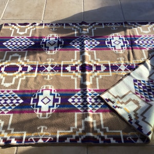 May include: A tan and cream-colored blanket with a geometric pattern. The design includes white, purple, blue, and yellow shapes. The blanket is folded over, revealing a cream-colored underside with a similar pattern.