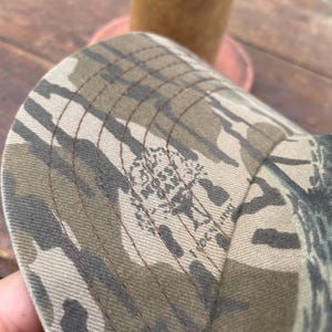Op de afbeelding: Een baseballpet met een camouflagepatroon in bruin-, beige- en donkergroene tinten. De stof is voorzien van een herhalend Mossy Oak boomlogo en is afgewerkt met zichtbare donkerbruine stiksels langs de panelen van de gebogen klep.