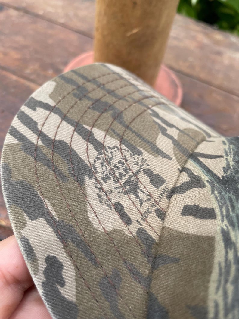 Op de afbeelding: Een baseballpet met een camouflagepatroon in bruin-, beige- en donkergroene tinten. De stof is voorzien van een herhalend Mossy Oak boomlogo en is afgewerkt met zichtbare donkerbruine stiksels langs de panelen van de gebogen klep.