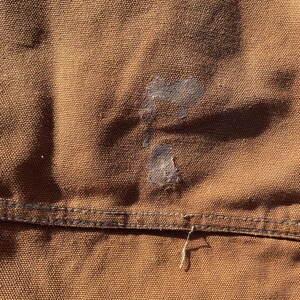 May include: Close-up of brown fabric with a visible stain and a seam. The fabric has a textured appearance, and the stitching is visible along the seam. A small, frayed thread hangs from the seam.