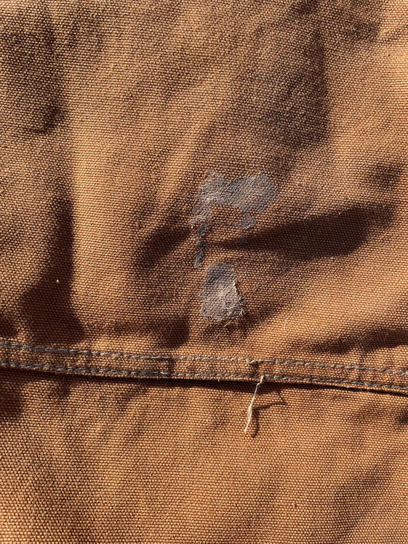 May include: Close-up of brown fabric with a visible stain and a seam. The fabric has a textured appearance, and the stitching is visible along the seam. A small, frayed thread hangs from the seam.