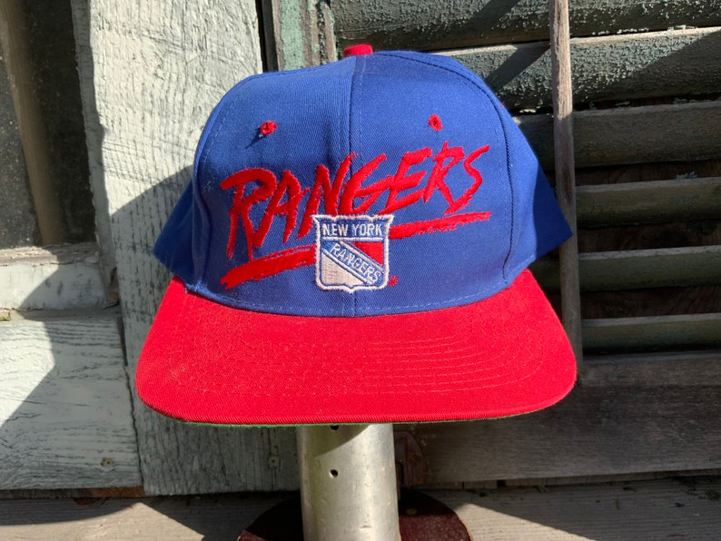 May include: A blue and red baseball cap with the words "New York Rangers" and a logo of the team on the front.