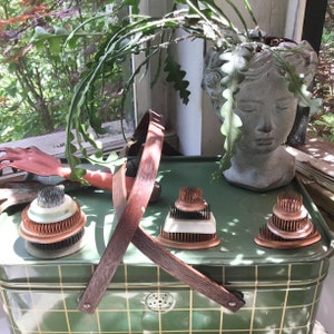 May include: A collection of floral arranging tools, including kenzans and a wooden stand, displayed on a green metal tray. The kenzans are copper and brass with various tiers. A stone head planter holds a trailing plant.