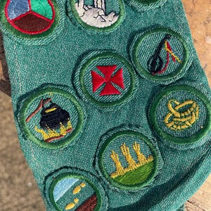 Op de afbeelding: Close-up van een groene stof met diverse geborduurde patches. De patches tonen verschillende ontwerpen, waaronder een rood kruis en een tent. De ronde patches met groene randen zijn waarschijnlijk gerelateerd aan scouting of een vergelijkbare organisatie.