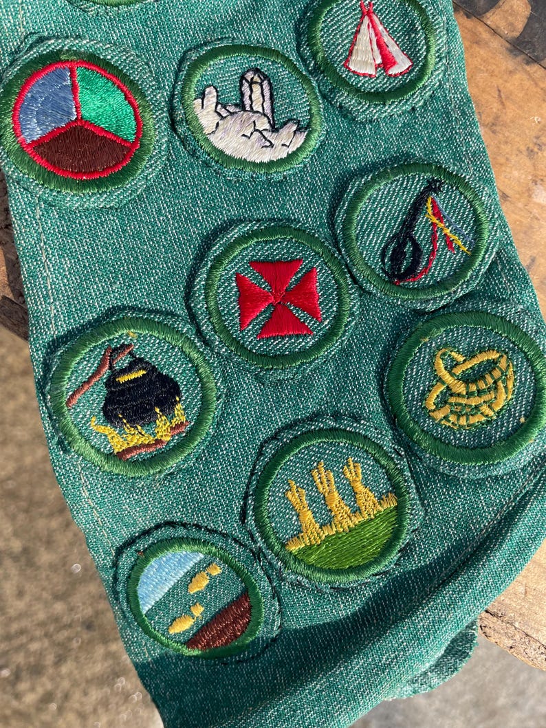 Op de afbeelding: Close-up van een groene stof met diverse geborduurde patches. De patches tonen verschillende ontwerpen, waaronder een rood kruis en een tent. De ronde patches met groene randen zijn waarschijnlijk gerelateerd aan scouting of een vergelijkbare organisatie.