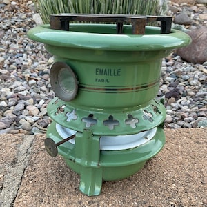 May include: A vintage green enamel stove with the text "EMAILLE FABR." The stove has a round shape with a metal grate on top and decorative cutouts. It is a classic piece of kitchenware.