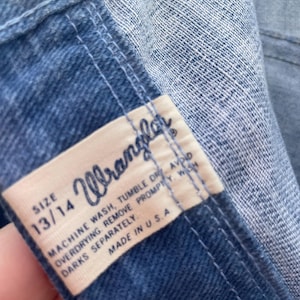 Pu&ograve; includere: Primo piano di una sezione di jeans blu. Un'etichetta rettangolare con il marchio "Wrangler" &egrave; visibile, insieme alla taglia 13/14. L'etichetta include anche le istruzioni di lavaggio e "Made in U.S.A."