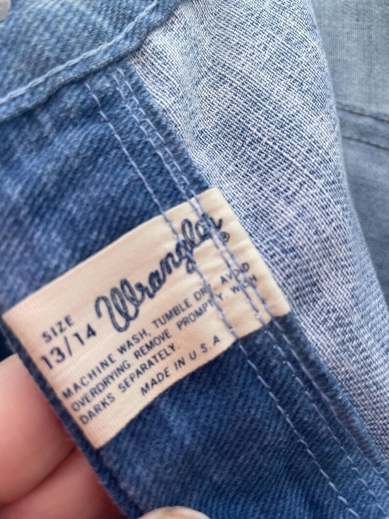 Pu&ograve; includere: Primo piano di una sezione di jeans blu. Un'etichetta rettangolare con il marchio "Wrangler" &egrave; visibile, insieme alla taglia 13/14. L'etichetta include anche le istruzioni di lavaggio e "Made in U.S.A."