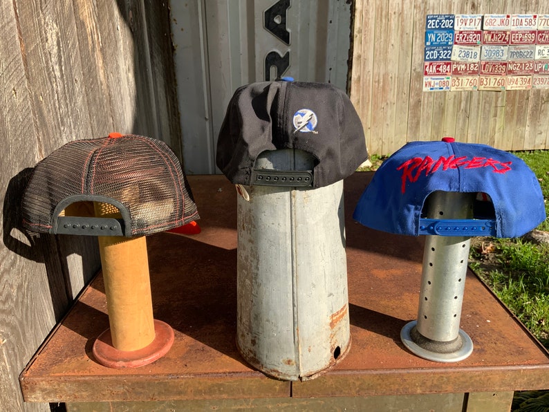 May include: Three baseball caps displayed on stands. The cap on the left is black with a red mesh back and a black brim. The middle cap is black with a blue lightning bolt logo. The cap on the right is blue with a red "RANGERS" logo.