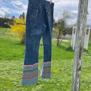 Pu&ograve; includere: Jeans in denim blu scuro con un motivo floreale decorativo sull'orlo svasato. I jeans sono appesi a un filo all'aperto. I jeans hanno un classico design a cinque tasche e un logo Wrangler.