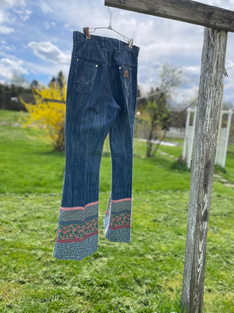 Pu&ograve; includere: Jeans in denim blu scuro con un motivo floreale decorativo sull'orlo svasato. I jeans sono appesi a un filo all'aperto. I jeans hanno un classico design a cinque tasche e un logo Wrangler.