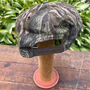 Op de afbeelding: Een baseballpet met een tijgerstreep camouflagepatroon in olijfgroen, bruin en zwart. De pet is voorzien van een bruine plastic snapback-sluiting aan de achterkant en kleine bruine metalen oogjes voor ventilatie op de zijpanelen.