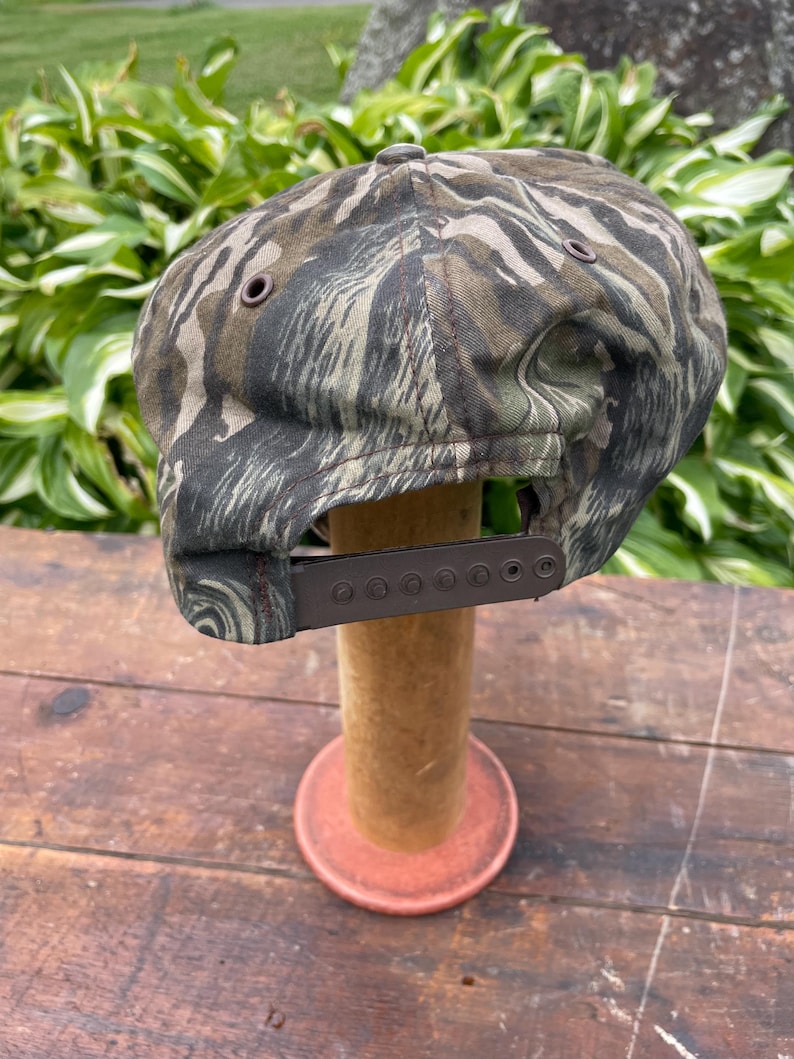 Op de afbeelding: Een baseballpet met een tijgerstreep camouflagepatroon in olijfgroen, bruin en zwart. De pet is voorzien van een bruine plastic snapback-sluiting aan de achterkant en kleine bruine metalen oogjes voor ventilatie op de zijpanelen.