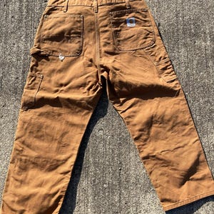 May include: Brown work pants with multiple pockets, including two back pockets and a side pocket. The pants have a straight leg design and a small logo patch on the back pocket. The pants are made of a durable, heavy-duty fabric.
