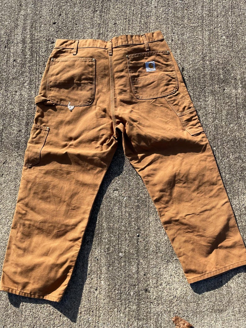 May include: Brown work pants with multiple pockets, including two back pockets and a side pocket. The pants have a straight leg design and a small logo patch on the back pocket. The pants are made of a durable, heavy-duty fabric.
